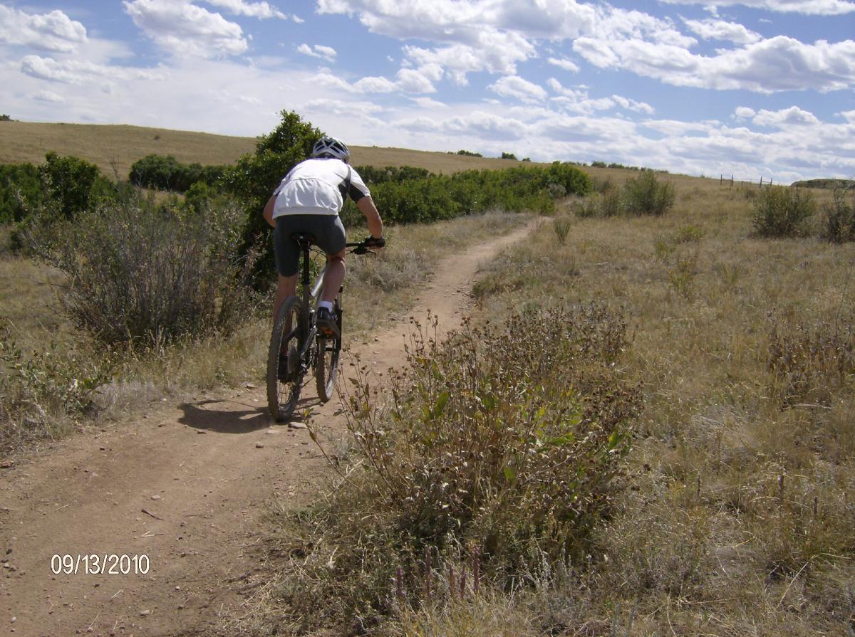 A person riding a mountain bike along a dirt trail in an open landscape, surrounded by grass and low bushes, under a partly cloudy sky. Ridgeline Open Space Trail mountain bike trail.