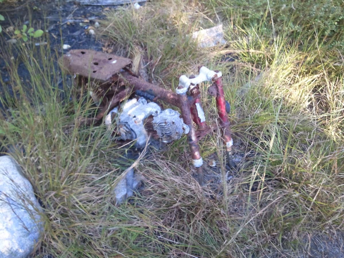 An old, rusted mechanical object partially submerged in grass and black mud, accompanied by remains of metal components. The scene includes rocks and overgrown foliage indicating a neglected area. Cutler Park mountain bike trail.