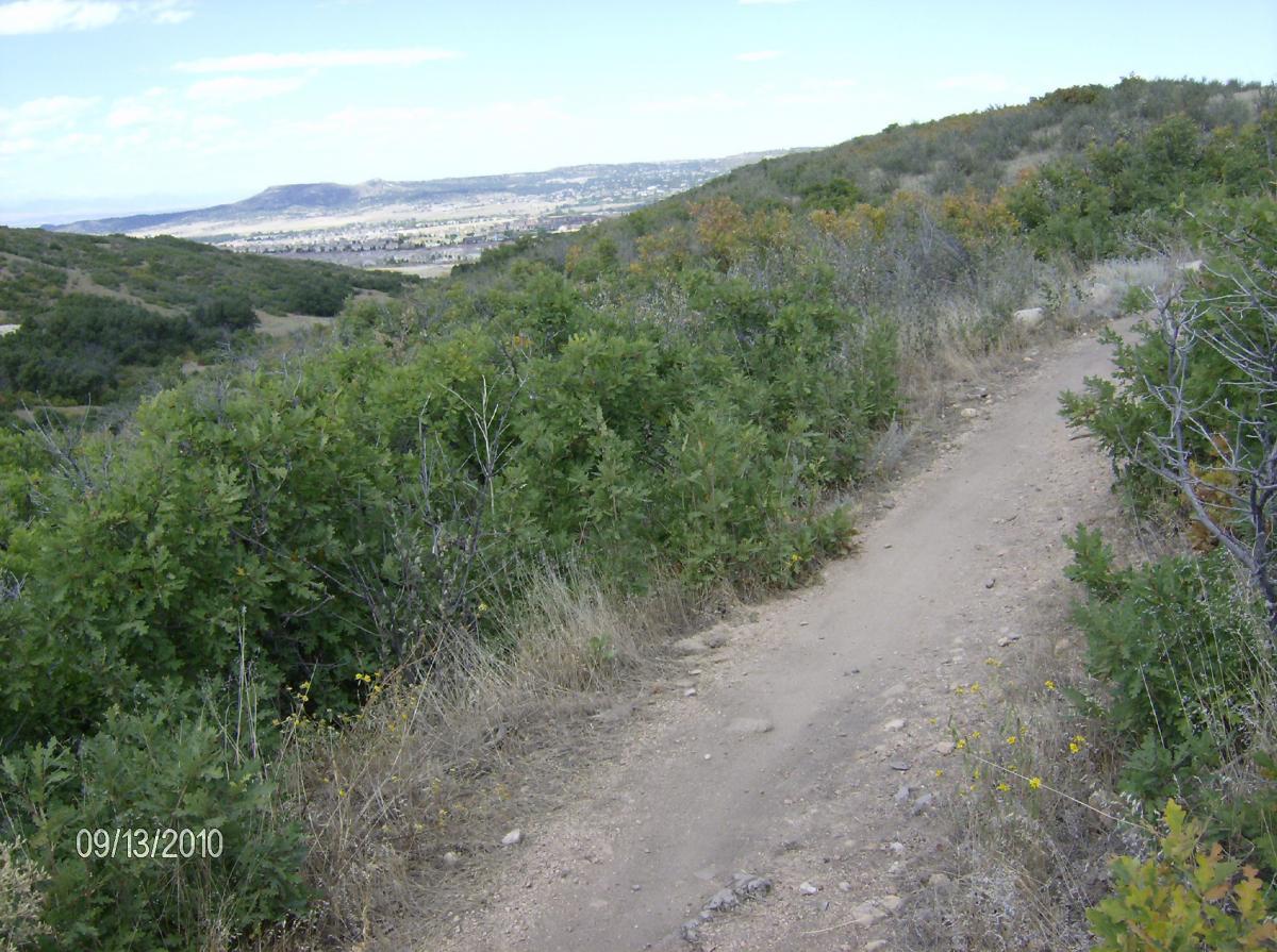 A dirt trail winds through a hilly landscape, surrounded by green bushes and sparse vegetation. The view overlooks a valley with distant hills and a partly cloudy sky. The date at the bottom indicates September 13, 2010. Ridgeline Open Space Trail mountain bike trail.