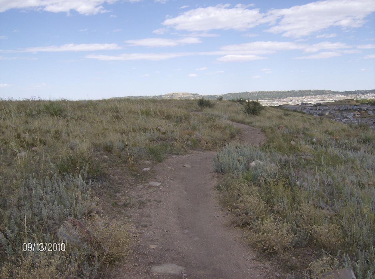 A dirt path winding through grassy terrain under a partly cloudy sky, with rolling hills and distant buildings visible in the background. The scene captures a natural landscape with sparse vegetation and a peaceful, outdoor atmosphere. Ridgeline Open Space Trail mountain bike trail.