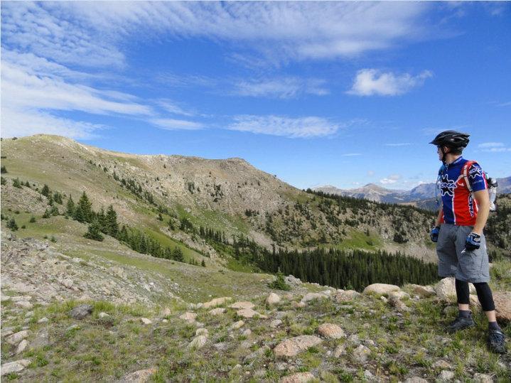 A person wearing a colorful cycling jersey and helmet stands on a rocky outcrop, gazing out over a scenic mountainous landscape with rolling hills and dense forests under a partly cloudy sky. Monarch Crest Trail mountain bike trail.