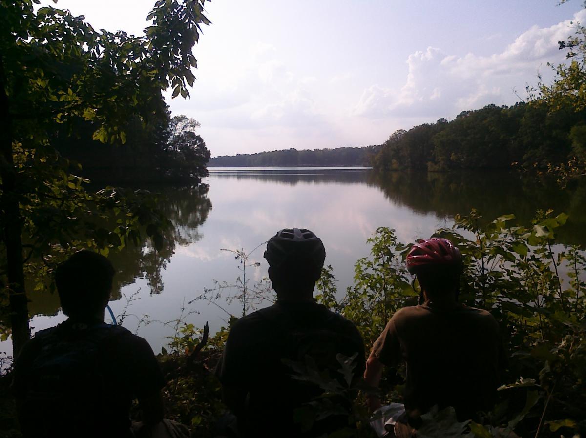 Three individuals wearing cycling helmets are sitting on the bank of a calm lake, surrounded by lush greenery. The water reflects the cloudy sky, creating a serene and peaceful atmosphere. Herb Parsons mountain bike trail.