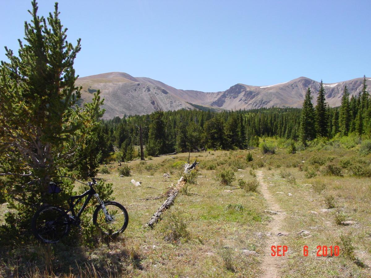 A scenic mountain landscape featuring a dirt biking trail surrounded by trees and lush greenery, with a bicycle leaning against a shrub in the foreground. The backdrop showcases rugged mountains under a clear blue sky. The image is dated September 6, 2010. Sheep Creek mountain bike trail.