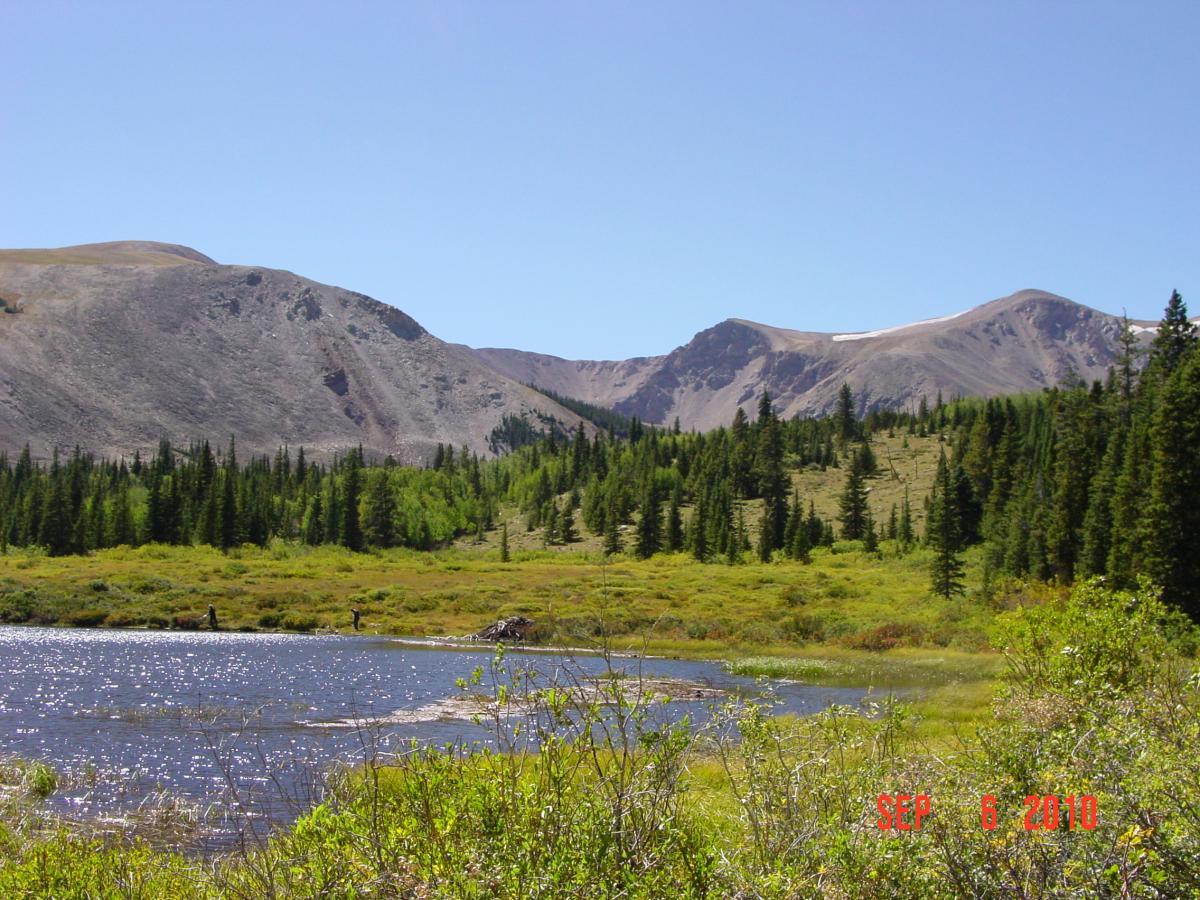 A scenic landscape featuring a serene lake in the foreground, surrounded by lush greenery and tall trees. In the background, rugged mountains rise under a clear blue sky, with some areas showing patches of snow. The image captures the beauty of nature in a tranquil setting. Sheep Creek mountain bike trail.