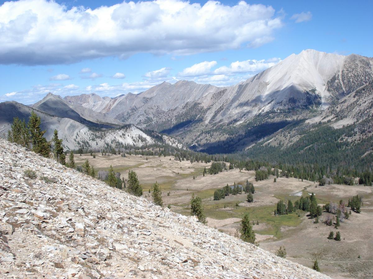 A panoramic view of a mountainous landscape featuring rugged peaks and rolling hills under a blue sky with scattered clouds. The foreground shows a rocky slope leading down to a valley with patches of greenery and trees, while the background highlights more distant mountain ranges. Fourth Of July Lake / Ants Basin / Robinson Bar mountain bike trail.