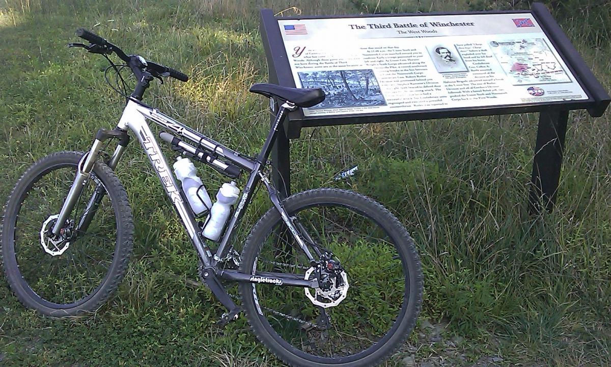 A mountain bike parked beside an informational sign about the Third Battle of Winchester, surrounded by grassy terrain. The sign features a historical overview and images related to the battle. 3rd Battle Of Winchester Trail mountain bike trail.