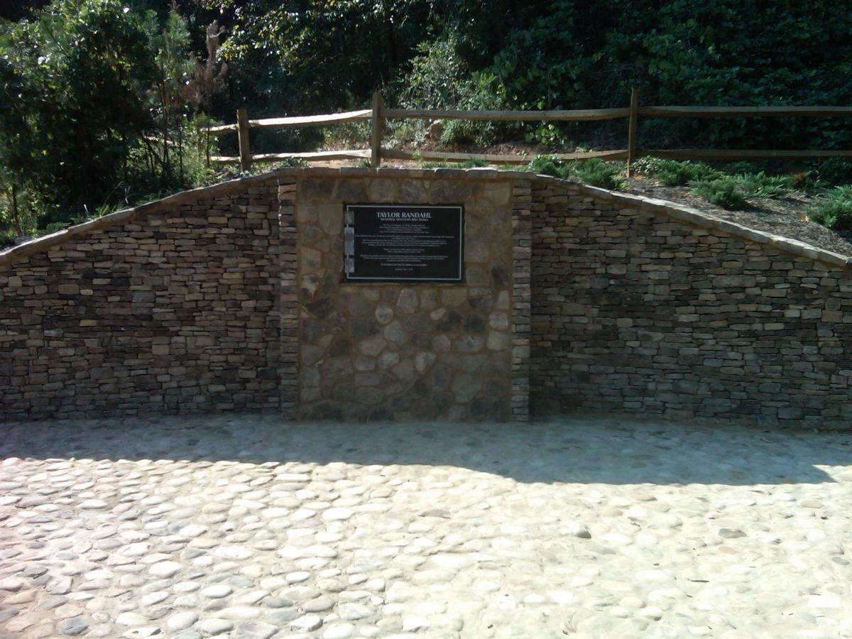 A stone wall with a plaque titled "Taylor Bandstand," surrounded by greenery. The area features a pathway made of cobblestones leading to the wall, which is built with a mix of small and large stones. A wooden fence is visible in the background, along with lush vegetation. Taylor Randahl Memorial Mountain Bike Trails At Olde Rope Mill Park mountain bike trail.