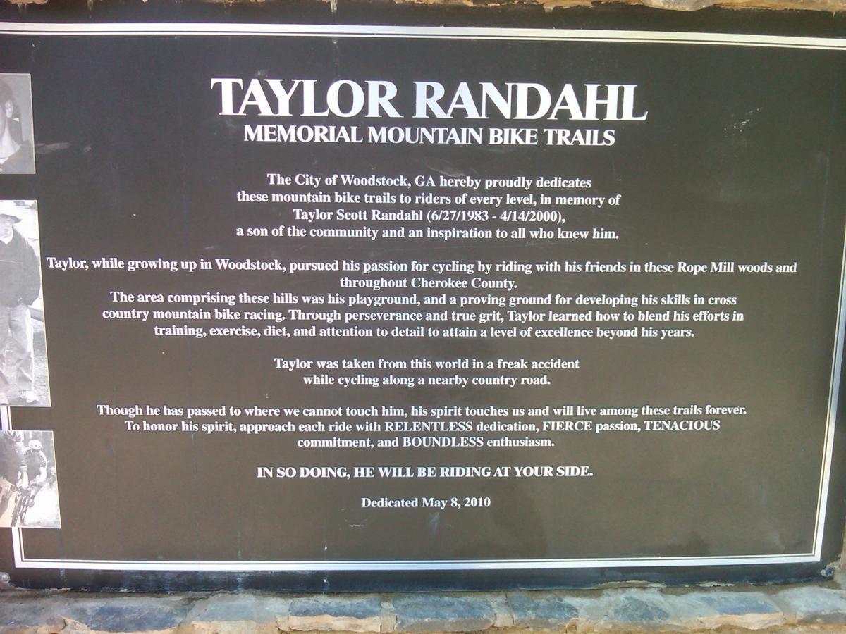 A black memorial plaque dedicated to Taylor Scott Randahl, featuring text honoring his passion for mountain biking and his legacy. The plaque includes his birth and death dates (6/27/1983 - 4/14/2000), a description of his dedication to cycling in Woodstock, GA, and a call to honor his spirit through commitment and enthusiasm while riding. Two black-and-white photographs of Taylor are placed on the left side. The plaque is set against a stone background and was dedicated on May 8, 2010. Taylor Randahl Memorial Mountain Bike Trails At Olde Rope Mill Park mountain bike trail.