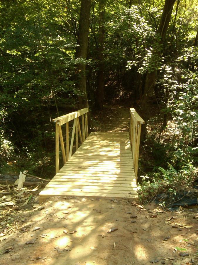 A wooden bridge leads over a small path in a dense forest, surrounded by lush greenery and sunlight filtering through the leaves. The ground is sandy with scattered leaves, and the scene evokes a sense of tranquility and nature. Taylor Randahl Memorial Mountain Bike Trails At Olde Rope Mill Park mountain bike trail.