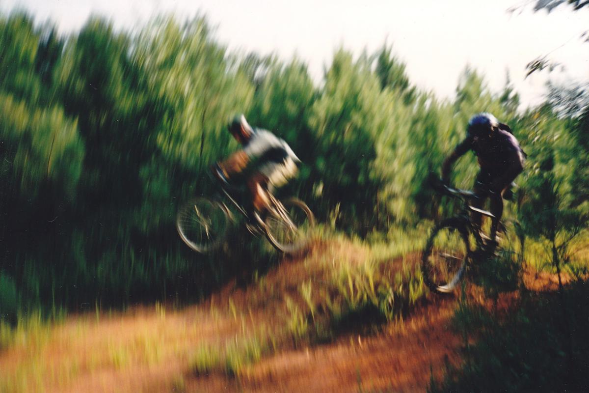 Two mountain bikers in motion, one performing a jump over a dirt mound, surrounded by lush green trees. The image has a slightly blurred effect, capturing the dynamic energy of mountain biking. Buncombe - Brickhouse Recreation Area mountain bike trail.