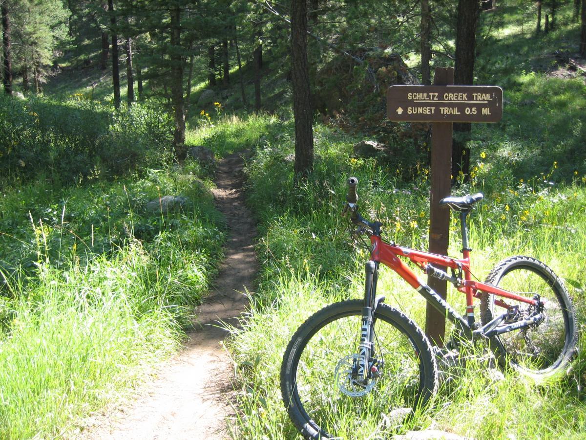 A mountain bike leaning against a wooden signpost that indicates the direction for the Schultz Creek Trail and Sunset Trail. The path is surrounded by lush green grass and trees in a sunny forested area. Schultz Creek mountain bike trail.
