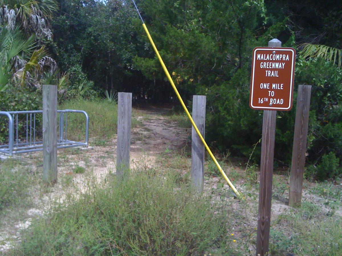 A pathway entrance to the Malacompra Greenway Trail, marked by a brown informational sign indicating "One Mile to 16th Road." The trail is bordered by wooden posts and features a bike rack in the foreground. Surrounding vegetation includes shrubs and small trees. Mala Compra mountain bike trail.