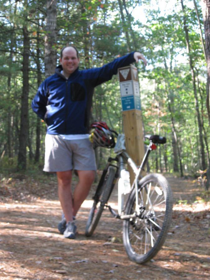 A person standing next to a trail marker in a wooded area, smiling while resting one hand on the marker and the other on a mountain bike. The scene captures a sunny day with trees in the background and a dirt trail leading into the forest. The person is wearing a blue jacket and shorts, with a bicycle helmet resting on the bike. Bear Bottom Loop mountain bike trail.