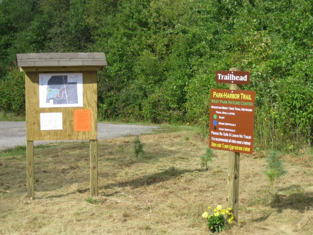 A wooden trailhead sign for the Park-Harbor Trail at West Park Nature Center, featuring a map and trail difficulty levels (Easy, More Difficult, Very Difficult). The sign encourages safety and helmet use for riders under 12 years old. The surrounding area is grassy with sparse vegetation. Park-harbor Trail mountain bike trail.