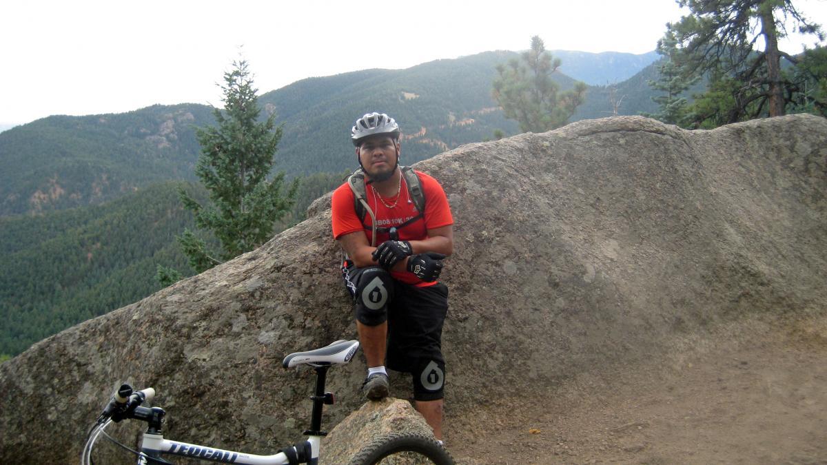 A mountain biker rests on a rock with a scenic view of rolling green hills in the background. He wears a red t-shirt, black shorts, and protective gear, including gloves and a helmet. A mountain bike is positioned beside him on a dirt path. Palmer Trail / Section 16 mountain bike trail.