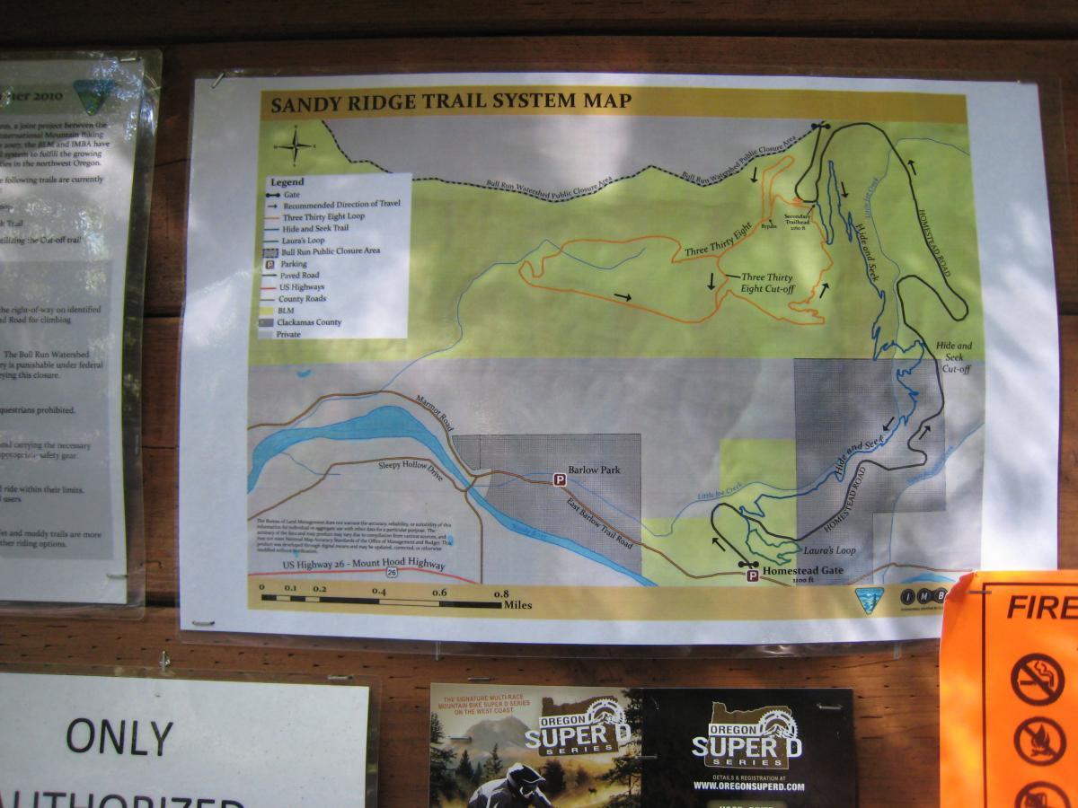 A trail system map for Sandy Ridge, showing various trails including the Three Thirty Eight Loop and Hide and Seek Trail, along with landmarks such as Barlow Park and parking areas. The map features a legend indicating different types of roads and trail usage, as well as a scale for distance measurement. Other nearby notices and information about the area are visible on the board. Sandy Ridge mountain bike trail.
