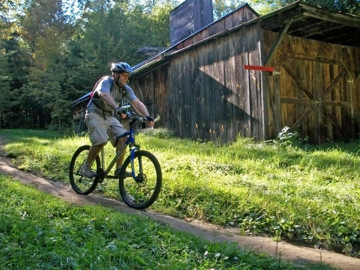 A person riding a mountain bike along a grassy trail in a wooded area, with a wooden barn in the background. The cyclist is wearing a helmet and casual cycling attire, enjoying the outdoor scenery. Kingdom Trails mountain bike trail.
