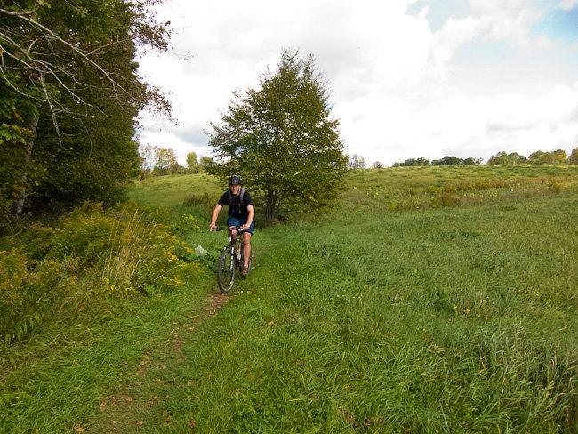A cyclist riding on a narrow dirt path through a green field, surrounded by trees and overcast sky. Kingdom Trails mountain bike trail.
