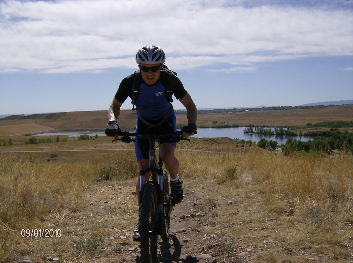 A mountain biker wearing a blue and black cycling outfit and a helmet is ascending a rocky trail with a scenic view of a river and rolling hills in the background under a clear blue sky. Bear Creek Lake Park mountain bike trail.