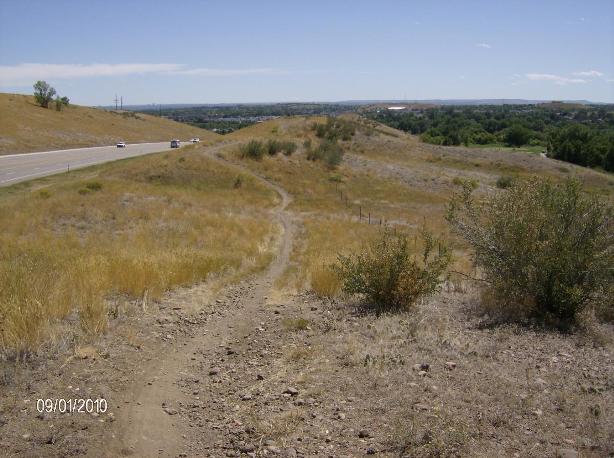A dirt path winding through dry, grassy hills, leading away from a highway. The landscape features sparse vegetation and a clear blue sky. In the distance, a small town can be seen among the hills. The date at the bottom shows September 1, 2010. Bear Creek Lake Park mountain bike trail.