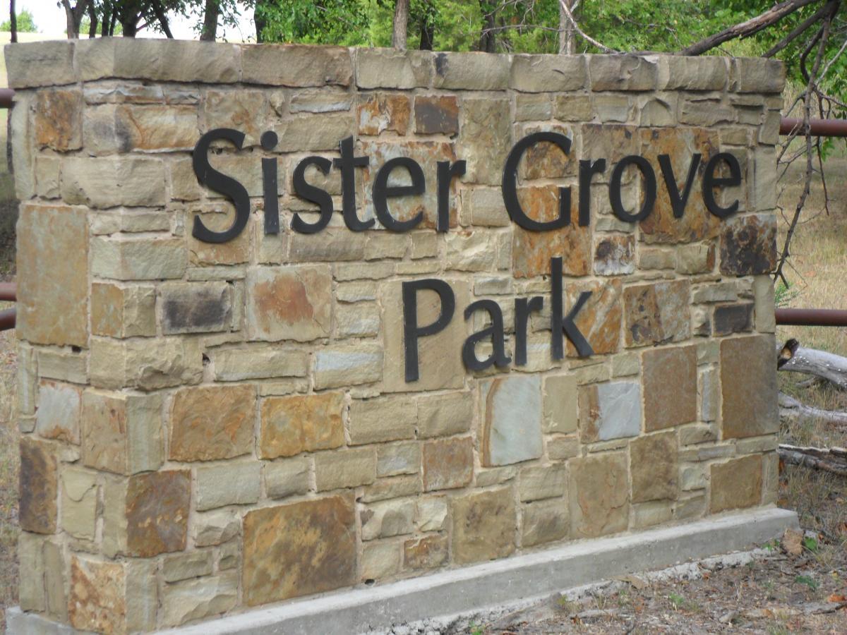 Stone sign for Sister Grove Park, featuring bold lettering against a textured stone background, surrounded by greenery. Sister Grove mountain bike trail.