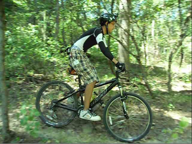 A young cyclist riding a mountain bike along a trail in a forested area, wearing a black and white cycling outfit and plaid shorts, with trees and greenery surrounding him. Chimney Rock mountain bike trail.