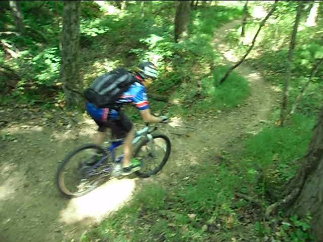 Mountain biker navigating a dirt trail in a green, wooded area, with sunlight filtering through the trees. The rider is wearing a helmet and a blue jersey, and the bike is positioned on a curved path. Chimney Rock mountain bike trail.