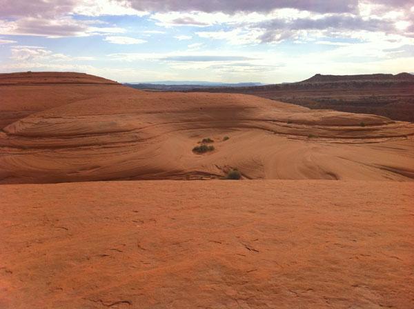 A panoramic view of a rugged, arid landscape featuring layered sandstone formations and gently sloping hills under a cloudy sky. Sparse vegetation can be seen in the foreground, contrasting with the vast expanse of red sand and rocky terrain. The distant horizon showcases more layered ridges and a dramatic sky. Bartlett Wash mountain bike trail.