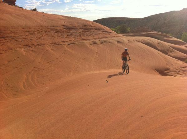 A person riding a mountain bike on smooth, sandy terrain with orange and reddish hues, surrounded by rolling hills and a clear blue sky in the background. Bartlett Wash mountain bike trail.