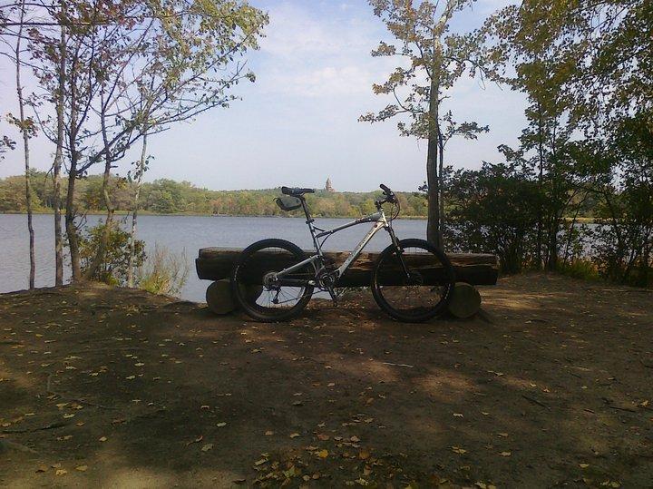 A mountain bike rests on a wooden bench by a serene lake, surrounded by trees and autumn leaves. The sky is partly cloudy, creating a peaceful outdoor scene. Cutler Park mountain bike trail.