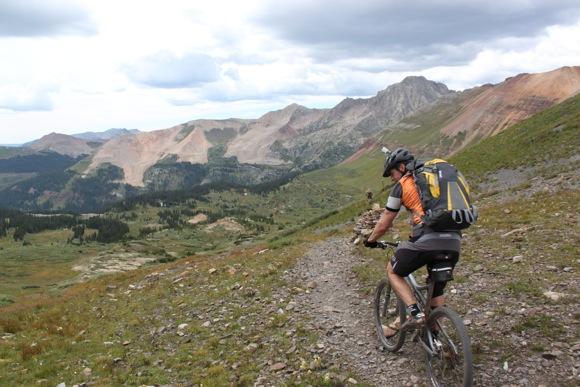 A mountain biker navigating a rocky trail on a mountainous landscape, surrounded by green valleys and rocky peaks under a cloudy sky. The cyclist is wearing a helmet and a backpack, showcasing the adventurous spirit of outdoor activities. Colorado Trail: Molas Pass To Bolam Pass Rd. mountain bike trail.