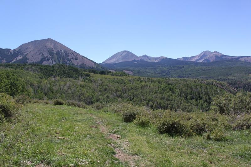 A panoramic view of a mountainous landscape under a clear blue sky, featuring several peaks in the distance. In the foreground, a lush green field with scattered shrubs and a dirt path leads into the scene. The surrounding mountains are characterized by varying heights and textures, showcasing a mix of forested areas and rocky terrain. Hazard County mountain bike trail.