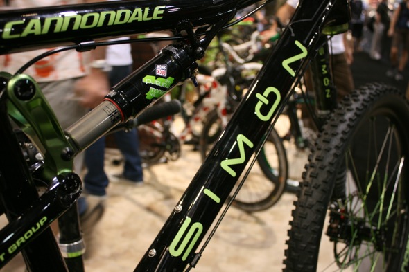 Close-up view of a Cannondale mountain bike frame featuring bold green and black lettering. The bike's shock absorber is visible, highlighting its suspension system. In the background, other bicycles and people are partially blurred, indicating a busy outdoor event or exhibition.