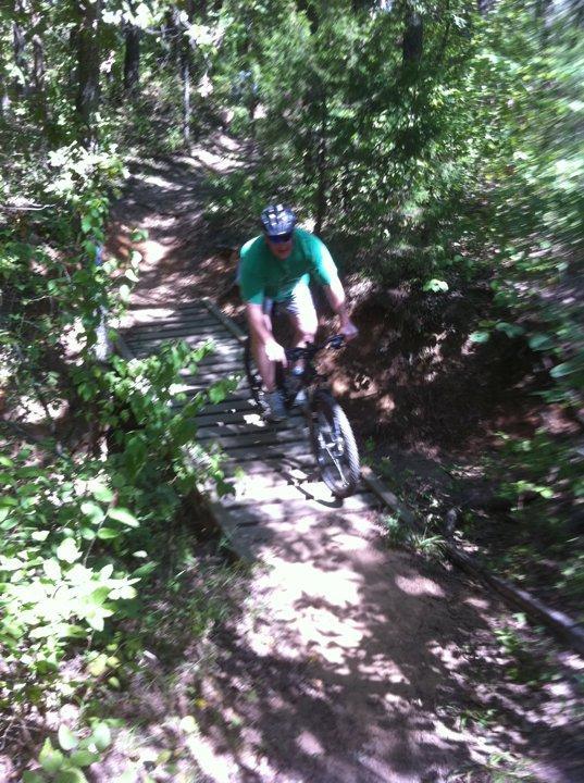 A person riding a mountain bike on a narrow wooden bridge in a wooded area. Sunlight filters through the trees, casting dappled shadows on the path. The rider is wearing a helmet and a green shirt, navigating the trail surrounded by greenery and dirt. McMurtry Trail mountain bike trail.