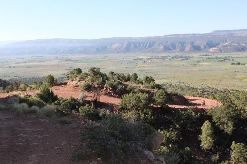 A scenic view of a rugged, rocky landscape, with a path winding through green and brown vegetation. In the background, rolling hills and mountains stretch against a clear blue sky, while a dirt road leads into the distant valley. The foreground features sparse shrubs and trees, enhancing the natural beauty of the area. Paradox Trail mountain bike trail.