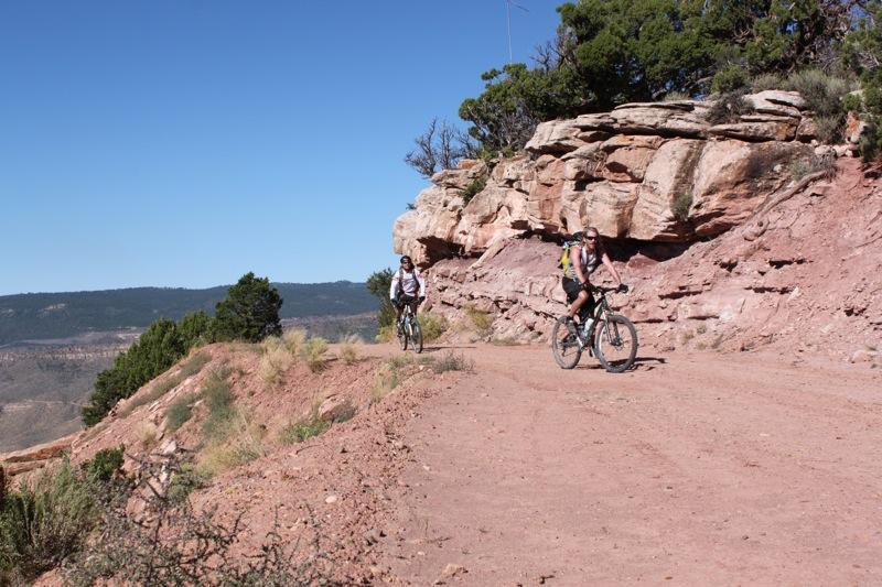 Two mountain bikers riding along a dirt trail that winds through red rock formations, surrounded by greenery and distant mountains under a clear blue sky. Paradox Trail mountain bike trail.