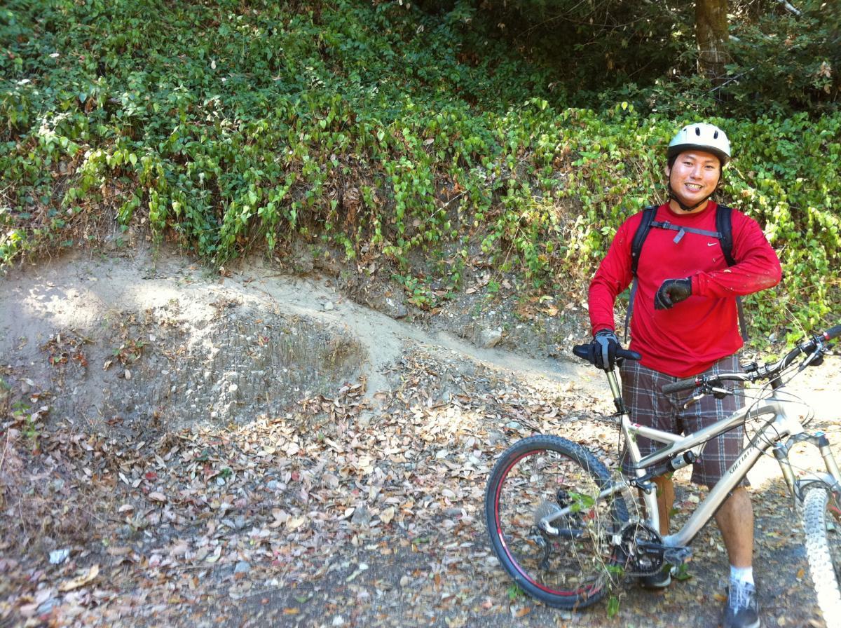 A person in a red long-sleeve shirt and helmet stands next to a mountain bike, smiling while resting on a dirt path surrounded by green foliage and fallen leaves. Stevens Canyon Trail mountain bike trail.