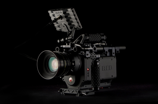 A professional cinema camera on a dark background, featuring a matte black design with a prominent lens, a rigged body, and various attachment points.