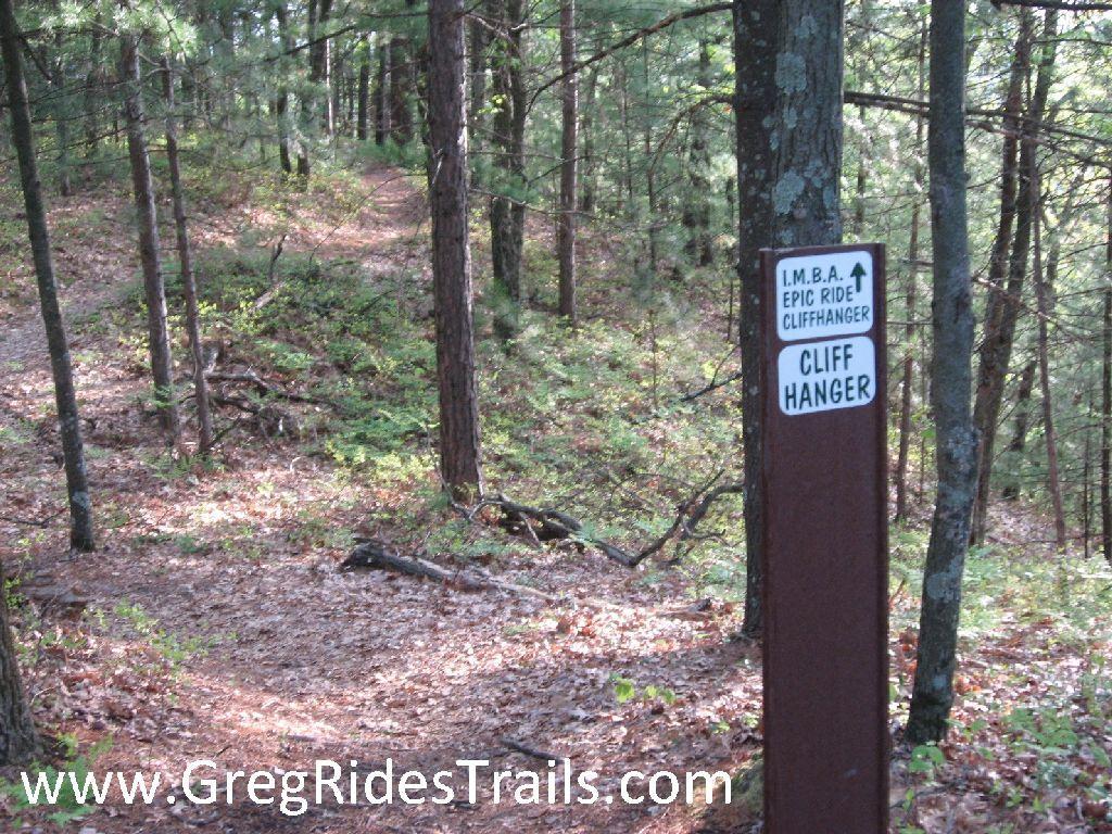 A forested trail intersection with a signpost indicating the "Cliff Hanger" trail, featuring markers for an epic ride as designated by IMBA (International Mountain Bicycling Association). The surrounding area is lush with greenery, and the path is covered in fallen leaves. Cliffhanger mountain bike trail.