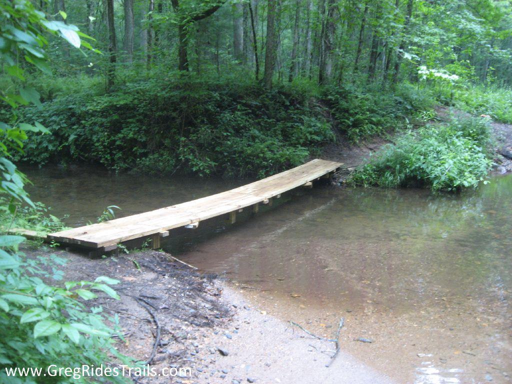 A wooden bridge crossing over a small stream in a lush green forest, surrounded by trees and ferns. The area shows a mix of sandy and earthy ground near the water's edge. Montgomery Creek Trail mountain bike trail.