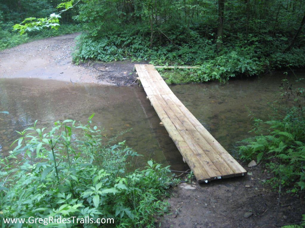 A wooden bridge crossing a small creek in a wooded area, surrounded by lush green foliage. A dirt path is visible leading towards the bridge, which offers a way to traverse the water. Montgomery Creek Trail mountain bike trail.