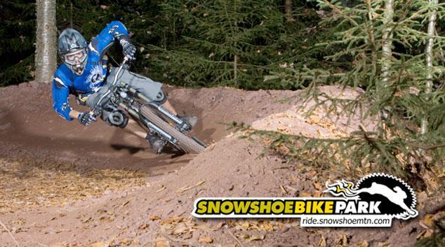 A mountain biker in a blue jersey leans into a turn on a dirt trail surrounded by trees, showcasing their skills and speed. The image includes the logo of Snowshoe Bike Park in the corner. Snowshoe Bike Park mountain bike trail.
