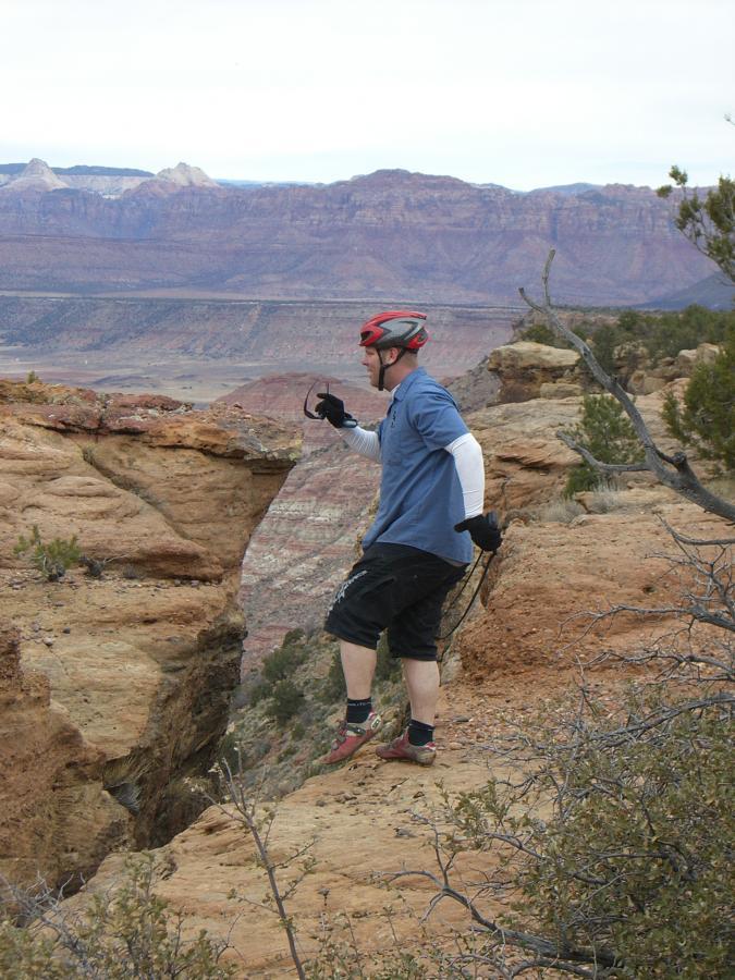 A person wearing a helmet and climbing gear is standing on a rocky ledge, overlooking a vast landscape with red rock formations and mountains in the background. The individual appears to be assessing the area, holding onto a climbing harness. The sky is overcast, adding a moody atmosphere to the scene. Gooseberry Mesa mountain bike trail.