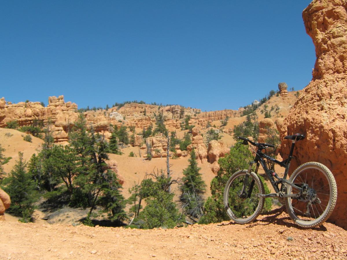 A mountain bike resting on a dirt trail surrounded by orange rock formations and green pine trees under a clear blue sky. The terrain features a mix of rugged hills and unique erosion patterns typical of a desert landscape. Thunder Mountain mountain bike trail.