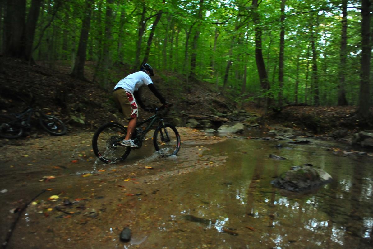 A person riding a mountain bike through a shallow stream in a wooded area, surrounded by greenery and trees. The cyclist is splashing water as they navigate over rocks and a sandy path. Several bicycles are visible in the background. Patapsco Valley State Park (Avalon Area) mountain bike trail.