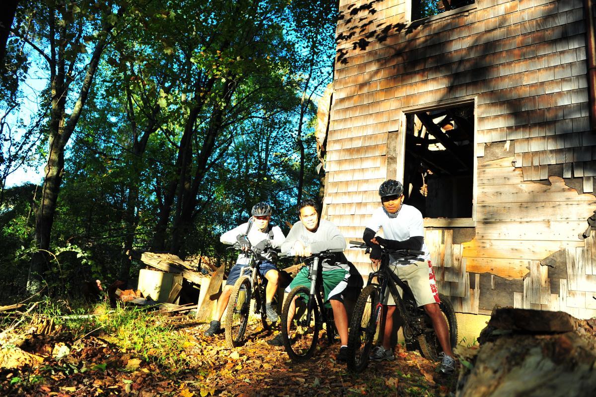 Three mountain bikers are gathered near an old, weathered building in a wooded area. The scene is set during daytime with sunlight filtering through the trees, casting a warm glow. Two of the bikers are wearing helmets and casual sportswear, while one is leaning on his bike looking intently at a device. The ground is covered with fallen leaves, and there are signs of disrepair around the building, adding to the adventurous atmosphere. Patapsco Valley State Park (Avalon Area) mountain bike trail.