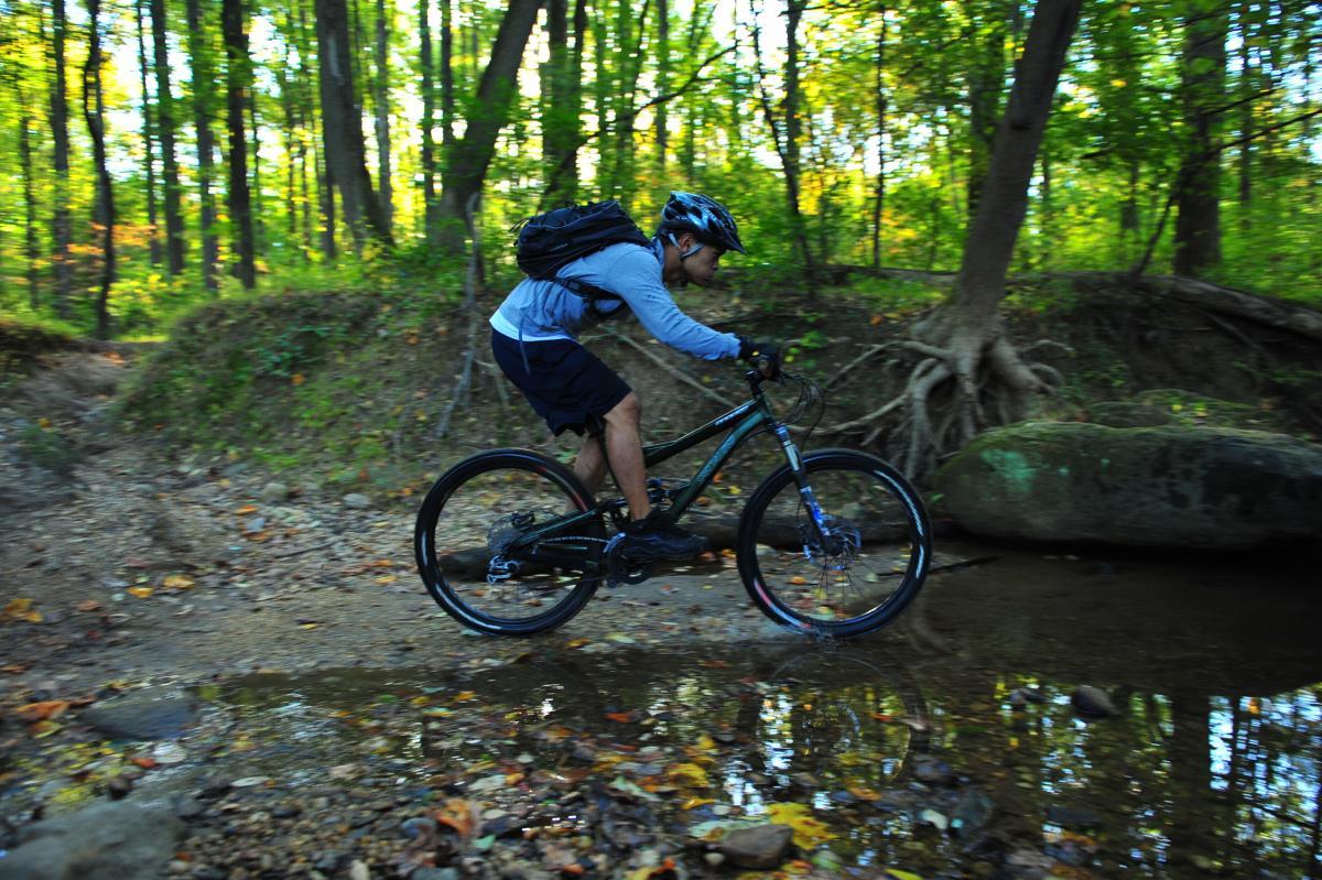 A person riding a mountain bike through a forested area, navigating over a rocky path beside a shallow stream. The scene features vibrant green trees and fallen leaves on the ground, capturing the essence of outdoor adventure. The cyclist is wearing a helmet and a backpack. Patapsco Valley State Park (Avalon Area) mountain bike trail.