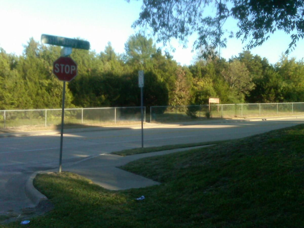A street corner featuring a stop sign and a street name sign, surrounded by greenery. The scene captures a clear sky and a paved road curving to the left, with a fence and trees in the background. Boulder Park mountain bike trail.