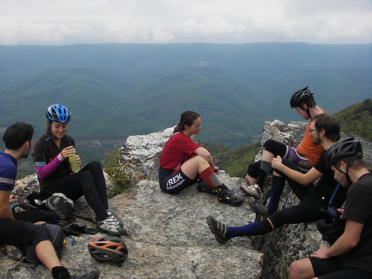 A group of six mountain bikers relaxing on a rocky ledge overlooking a scenic valley. They are dressed in cycling gear and helmets, enjoying a break while sharing drinks and conversation. The landscape features green hills and mountains under a cloudy sky. North Fork Mountain Trail mountain bike trail.