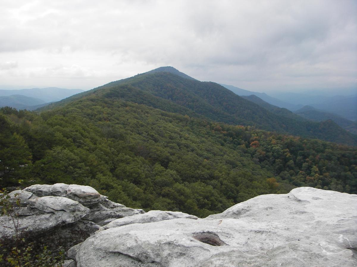 A panoramic view of a mountainous landscape featuring rolling hills blanketed with dense green forests. In the foreground, large gray rock formations are visible, leading to a ridge that showcases distant peaks under a cloudy sky. The scene conveys a sense of tranquility and natural beauty. North Fork Mountain Trail mountain bike trail.